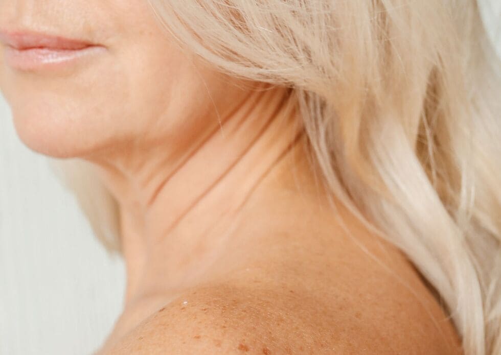 Plano detalle de cuello, hombro y media cara de una mujer madura con piel luminosa, símbolo de cuidado y bienestar.

